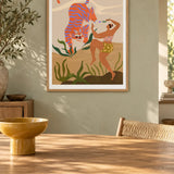 Dance With Me by Arty Guava – orange tiger with violet stripes beside a dancing woman and green leaves. Oak-framed print above a dining table in a sunlit sage room.