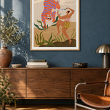 Dance With Me by Arty Guava – orange tiger with violet stripes beside a dancing woman and green leaves. Oak-framed print above a walnut sideboard against a blue living-room wall.
