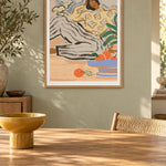 Lazy Days by Arty Guava – reclining woman in a floral blouse beside a blue bowl of oranges. Oak-framed print above a dining table in a sunlit sage room.