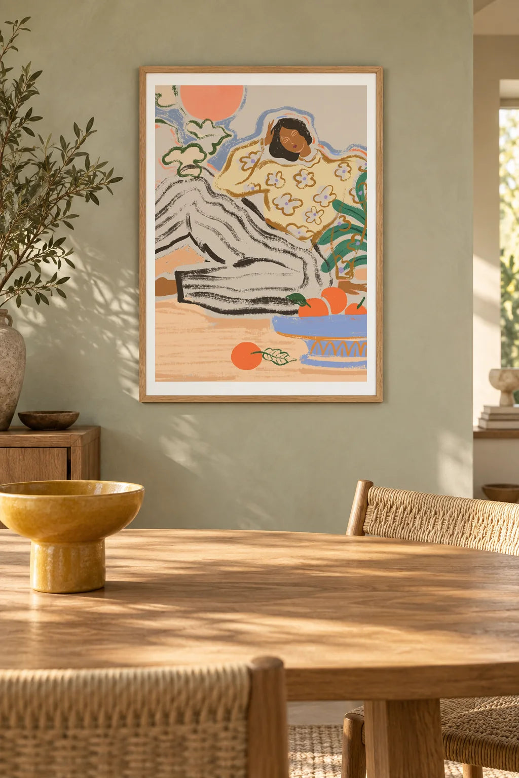 Lazy Days by Arty Guava – reclining woman in a floral blouse beside a blue bowl of oranges. Oak-framed print above a dining table in a sunlit sage room.