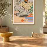 Lazy Days by Arty Guava – reclining woman in a floral blouse beside a blue bowl of oranges. Oak-framed print above a dining table in a sunlit sage room.