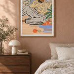 Lazy Days by Arty Guava – reclining woman in a floral blouse beside a blue bowl of oranges. Oak-framed print styled above a walnut bedside chest in a warm bedroom.