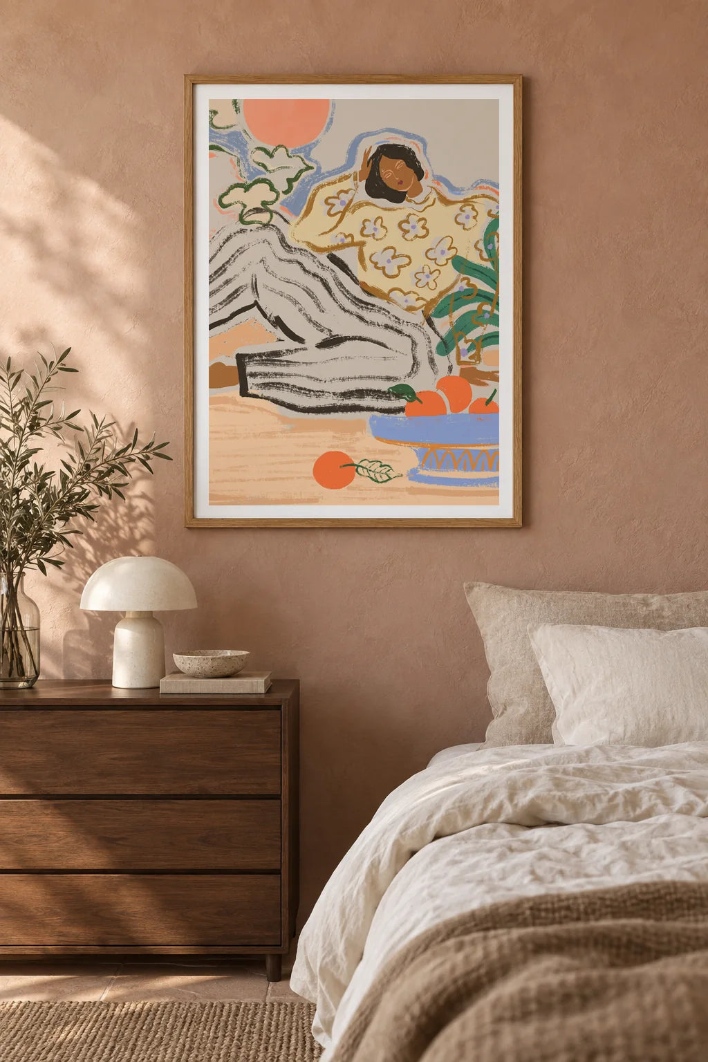 Lazy Days by Arty Guava – reclining woman in a floral blouse beside a blue bowl of oranges. Oak-framed print styled above a walnut bedside chest in a warm bedroom.
