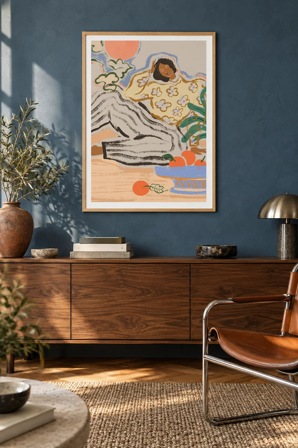Lazy Days by Arty Guava – reclining woman in a floral blouse beside a blue bowl of oranges. Oak-framed print above a walnut sideboard against a blue living-room wall.