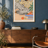 Lazy Days by Arty Guava – reclining woman in a floral blouse beside a blue bowl of oranges. Oak-framed print above a walnut sideboard against a blue living-room wall.