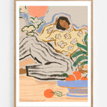 Lazy Days by Arty Guava – reclining woman in a floral blouse beside a blue bowl of oranges. Art print shown in a natural oak frame on a white wall.