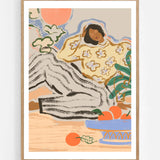 Lazy Days by Arty Guava – reclining woman in a floral blouse beside a blue bowl of oranges. Art print shown in a natural oak frame on a white wall.