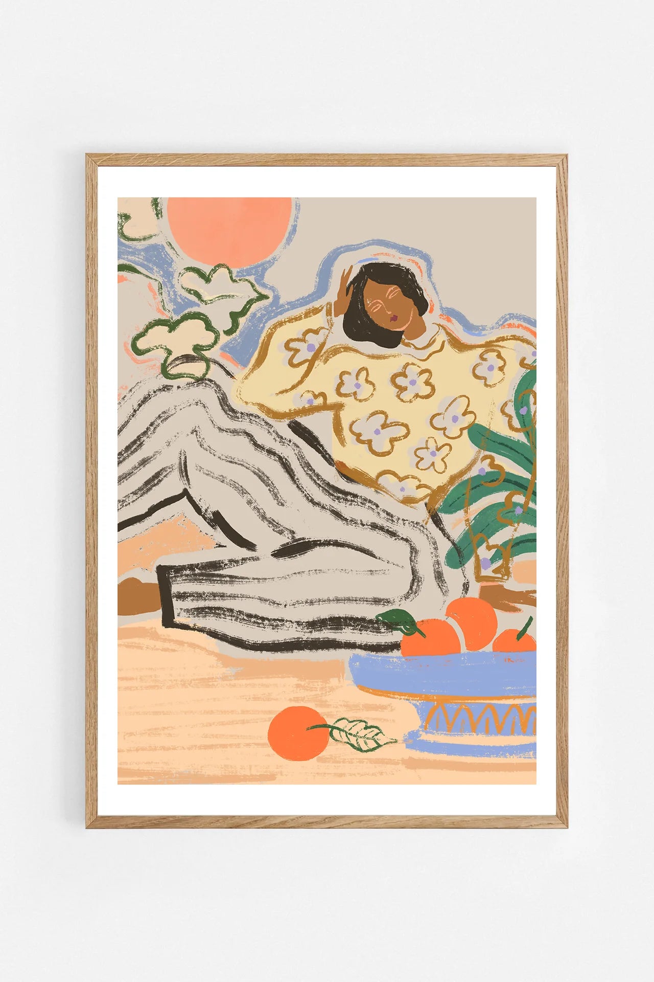 Lazy Days by Arty Guava – reclining woman in a floral blouse beside a blue bowl of oranges. Art print shown in a natural oak frame on a white wall.