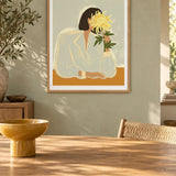 The Chrysanthemum by Arty Guava – woman holding a yellow chrysanthemum against a pale sage background. Oak-framed print above a dining table in a sunlit sage room.