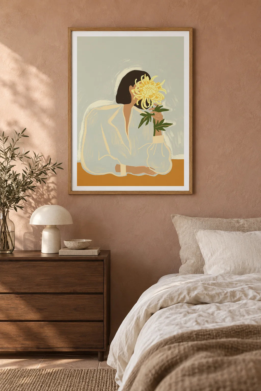 The Chrysanthemum by Arty Guava – woman holding a yellow chrysanthemum against a pale sage background. Oak-framed print styled above a walnut bedside chest in a warm bedroom.