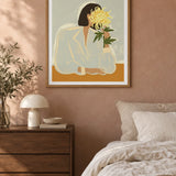 The Chrysanthemum by Arty Guava – woman holding a yellow chrysanthemum against a pale sage background. Oak-framed print styled above a walnut bedside chest in a warm bedroom.