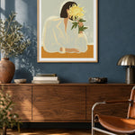 The Chrysanthemum by Arty Guava – woman holding a yellow chrysanthemum against a pale sage background. Oak-framed print above a walnut sideboard against a blue living-room wall.