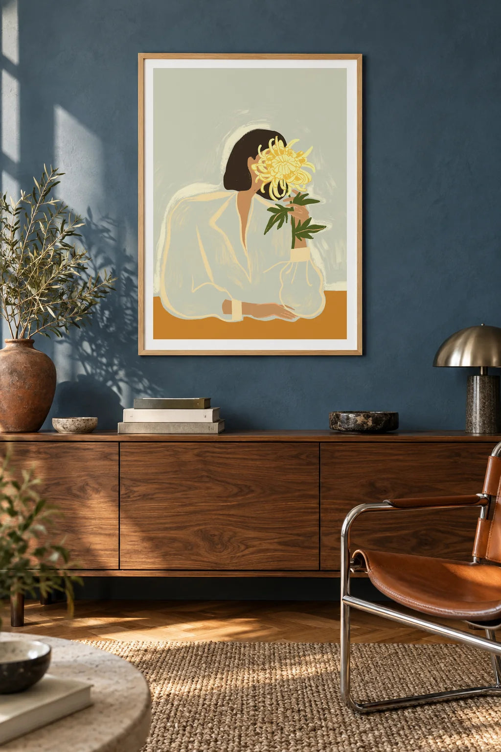 The Chrysanthemum by Arty Guava – woman holding a yellow chrysanthemum against a pale sage background. Oak-framed print above a walnut sideboard against a blue living-room wall.