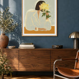 The Chrysanthemum by Arty Guava – woman holding a yellow chrysanthemum against a pale sage background. Oak-framed print above a walnut sideboard against a blue living-room wall.