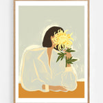 The Chrysanthemum by Arty Guava – woman holding a yellow chrysanthemum against a pale sage background. Art print shown in a natural oak frame on a white wall.