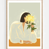 The Chrysanthemum by Arty Guava – woman holding a yellow chrysanthemum against a pale sage background. Art print shown in a natural oak frame on a white wall.