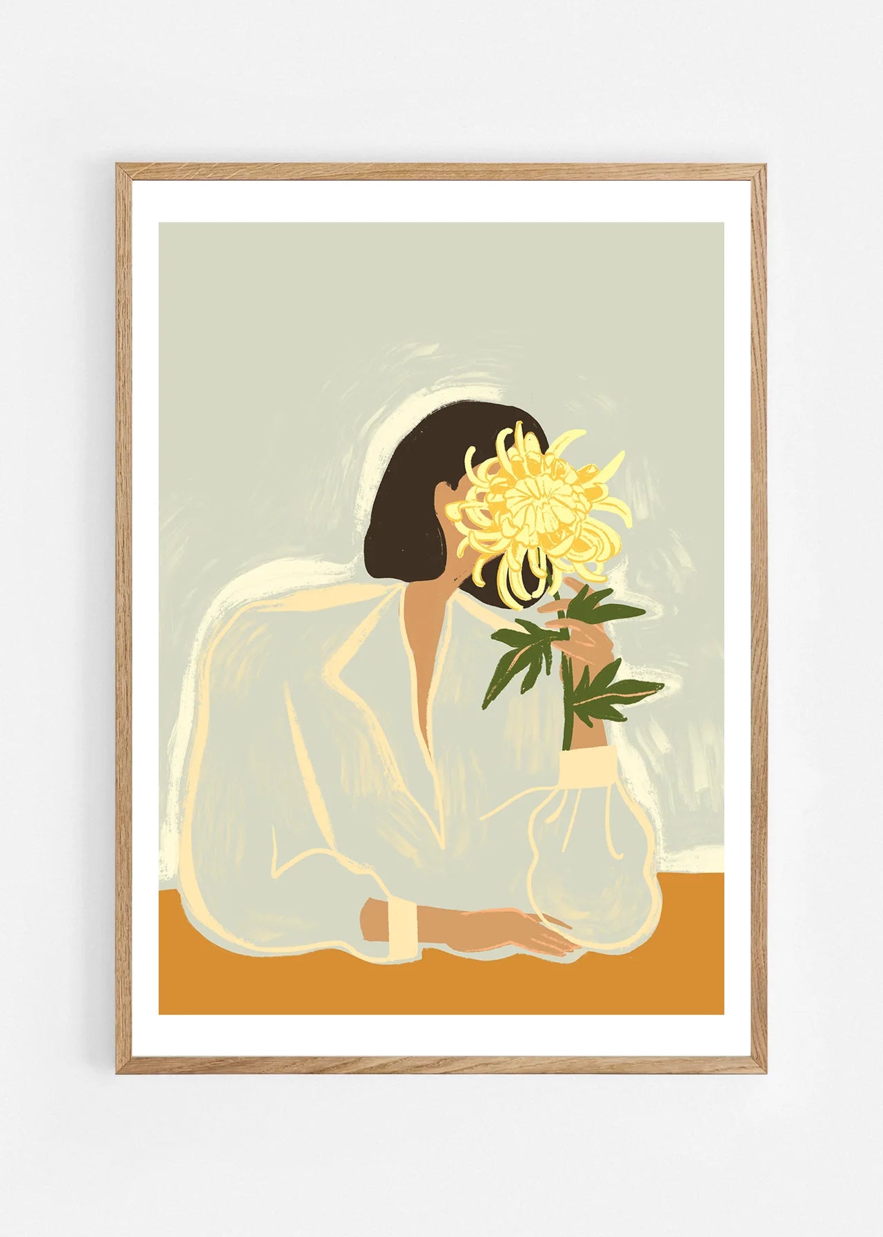 The Chrysanthemum by Arty Guava – woman holding a yellow chrysanthemum against a pale sage background. Art print shown in a natural oak frame on a white wall.