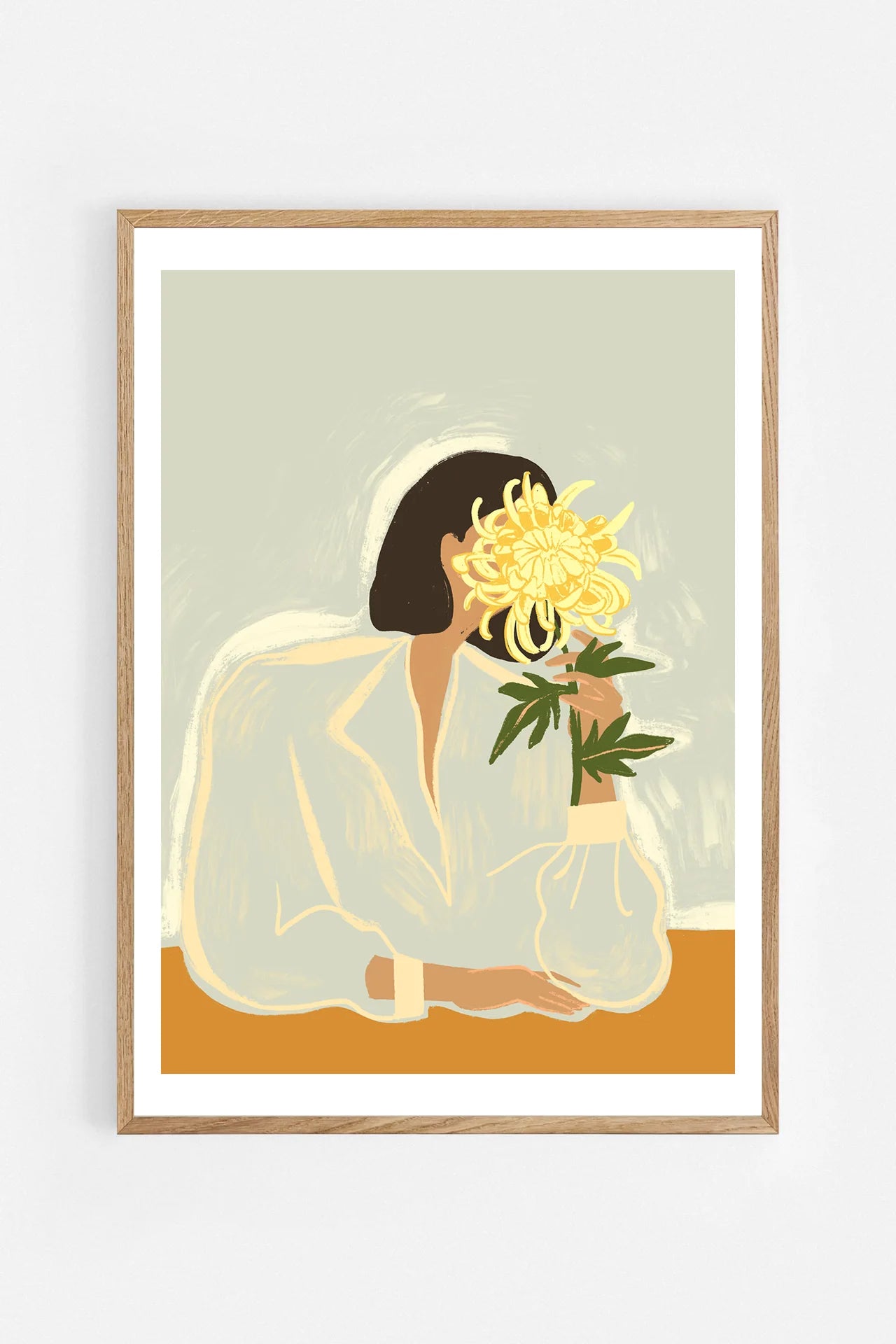 The Chrysanthemum by Arty Guava – woman holding a yellow chrysanthemum against a pale sage background. Art print shown in a natural oak frame on a white wall.